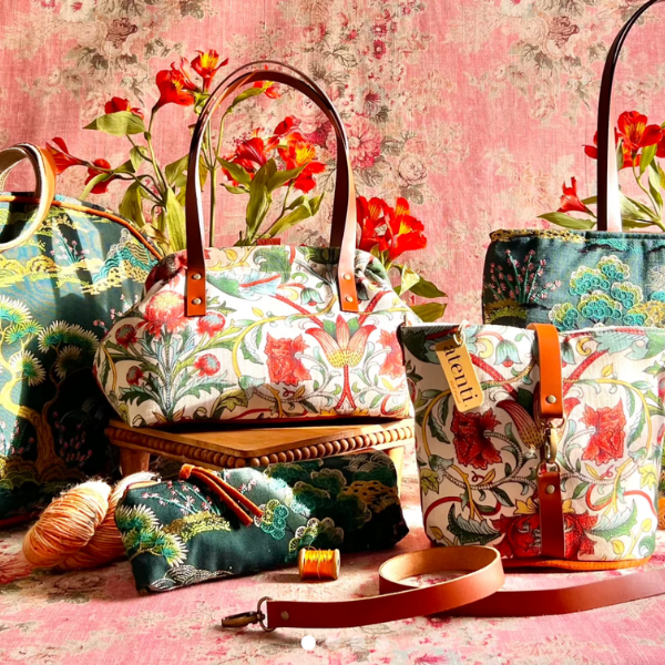 A styled product display of multiple handbags and a small pouch from the brand Kitchell, featuring a distinctive floral and botanical pattern in teal, red, and cream, with brown leather handles and straps, set against a pink floral wallpaper background.